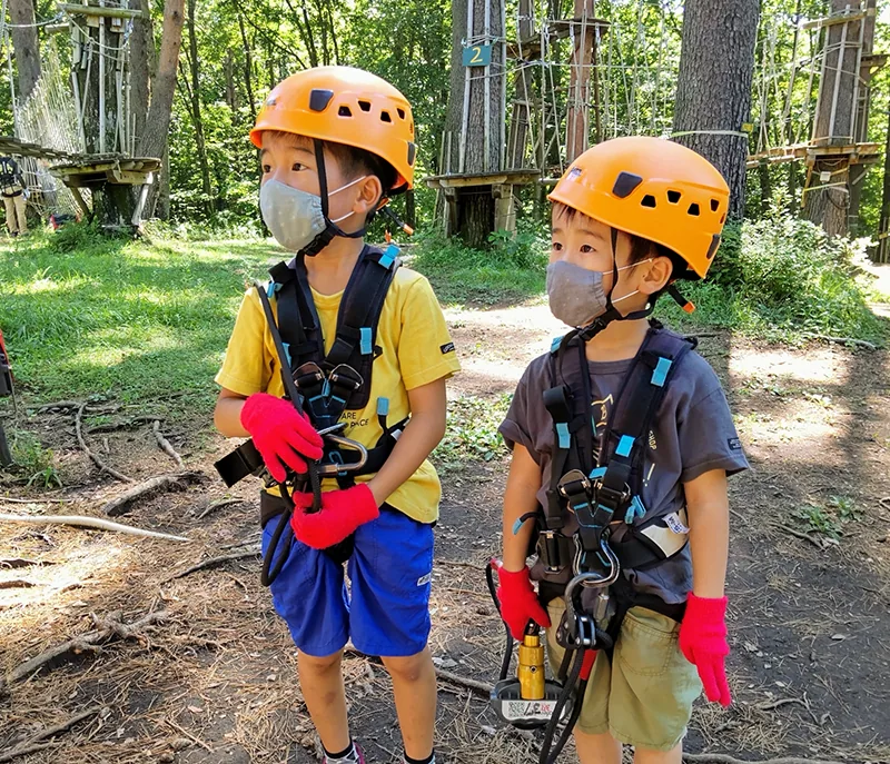 オレンジのヘルメットをかぶり森の中でアクティビティをする子供2人